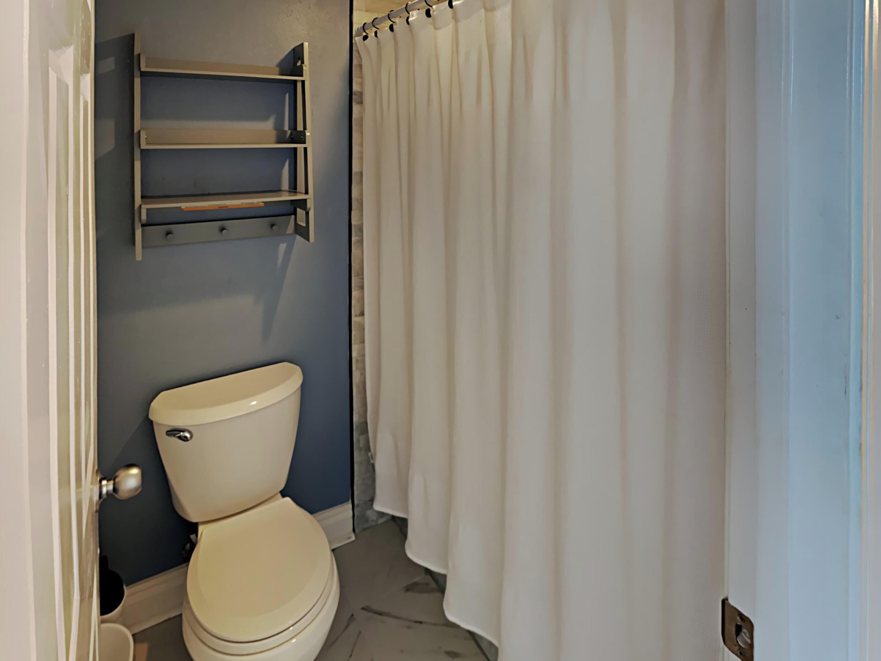 A bathroom with a toilet, a shower curtain, and a shelf above the toilet.