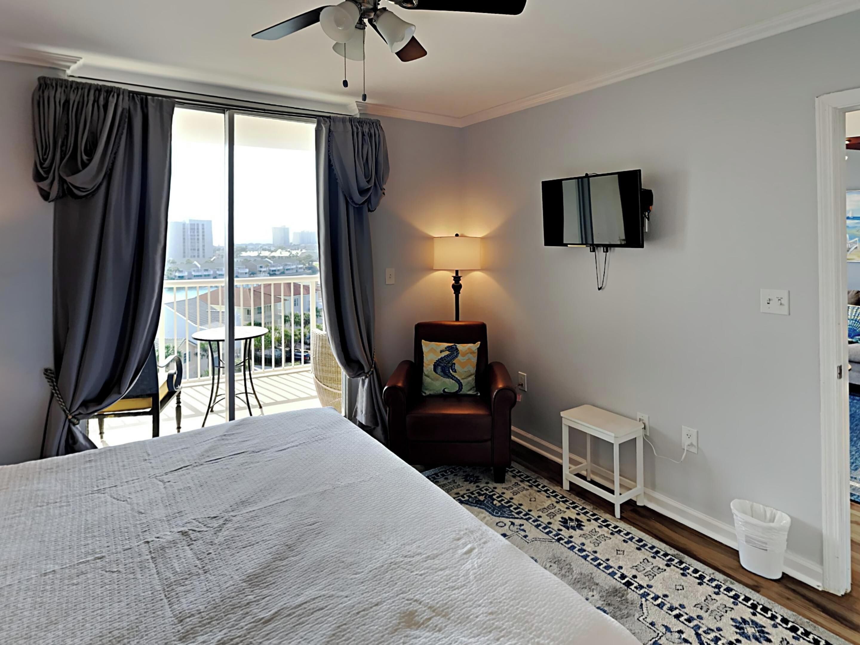 A hotel room with a large bed, a chair with a decorative pillow, a small table, a TV mounted on the wall, and a balcony with a view of the city.