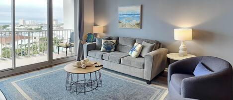 Een moderne woonkamer met een grijze bank, een ronde salontafel en een blauw accentkleed. Een plafondventilator en een schilderij zijn op de muur te zien.