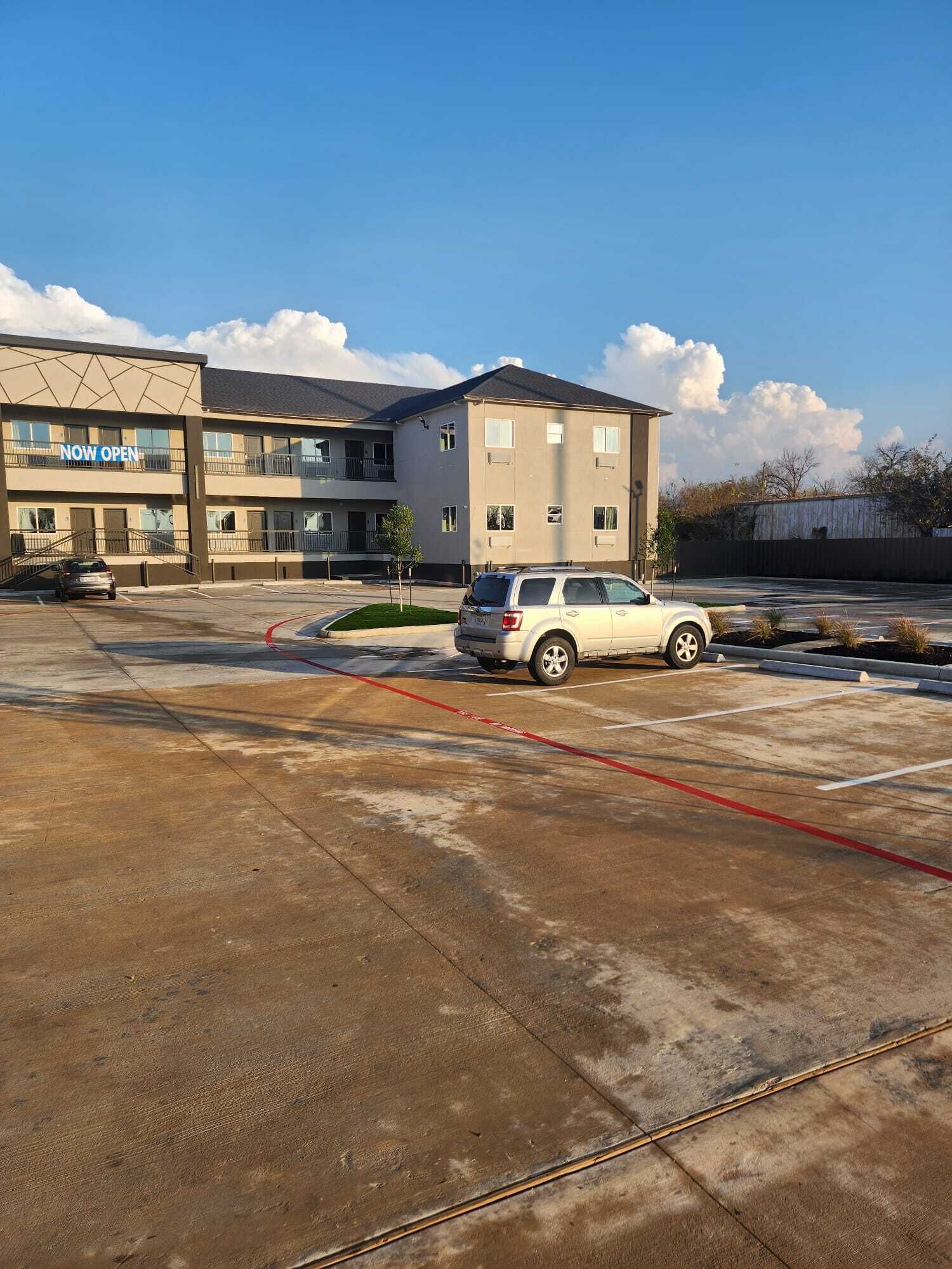 A parking lot with a white SUV, a building with a 'NOW OPEN' sign, and a clear blue sky.