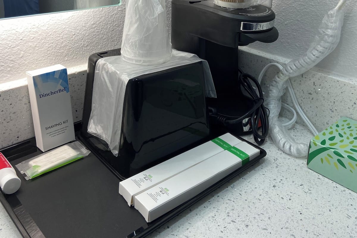 Uma bancada de banheiro com kit de barbear, máquina de café e dispensador de lenços.