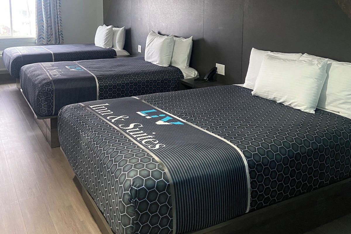 Quarto de hotel com três camas, cada uma com um cobreleja estampado escuro e almofadas brancas. O quarto tem um design moderno, com cabeceira escura e uma janela com cortinas.