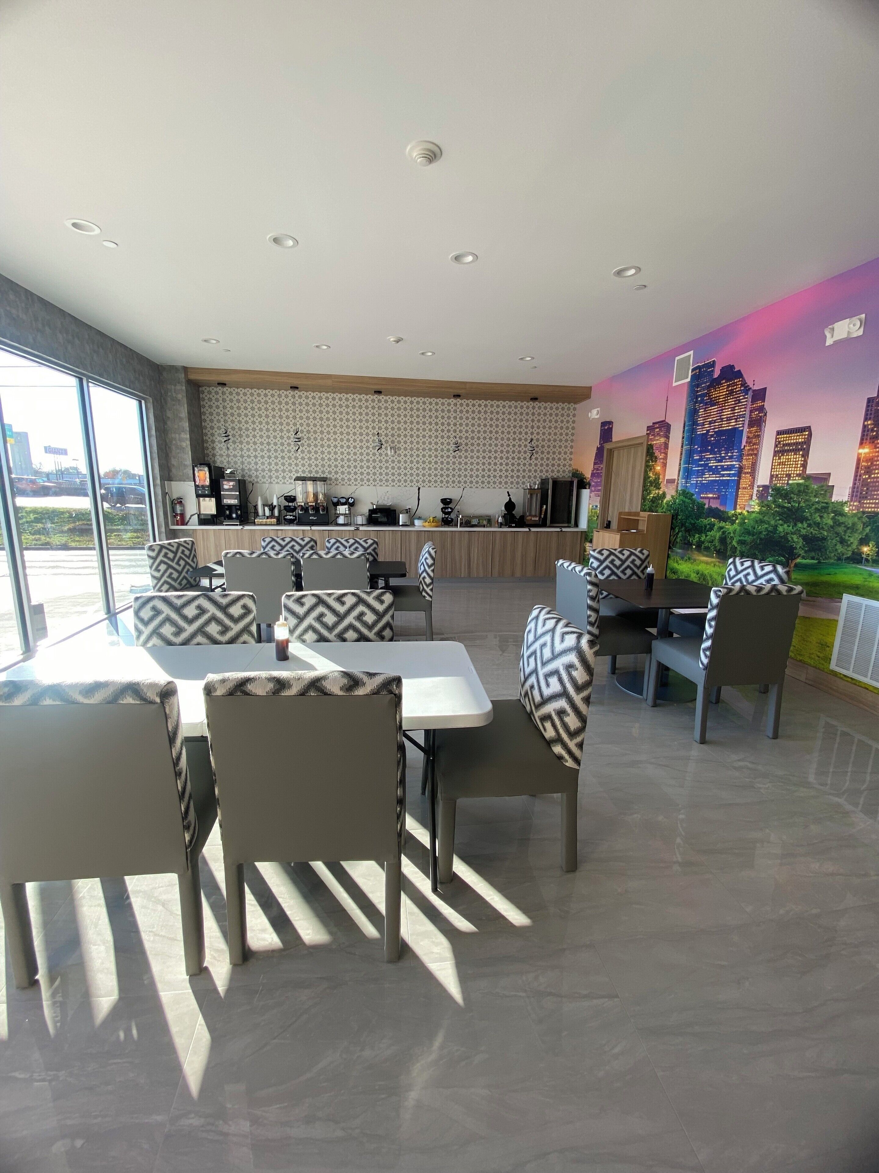 A modern dining area with patterned chairs and tables, a kitchen in the background, and a cityscape mural on the wall.