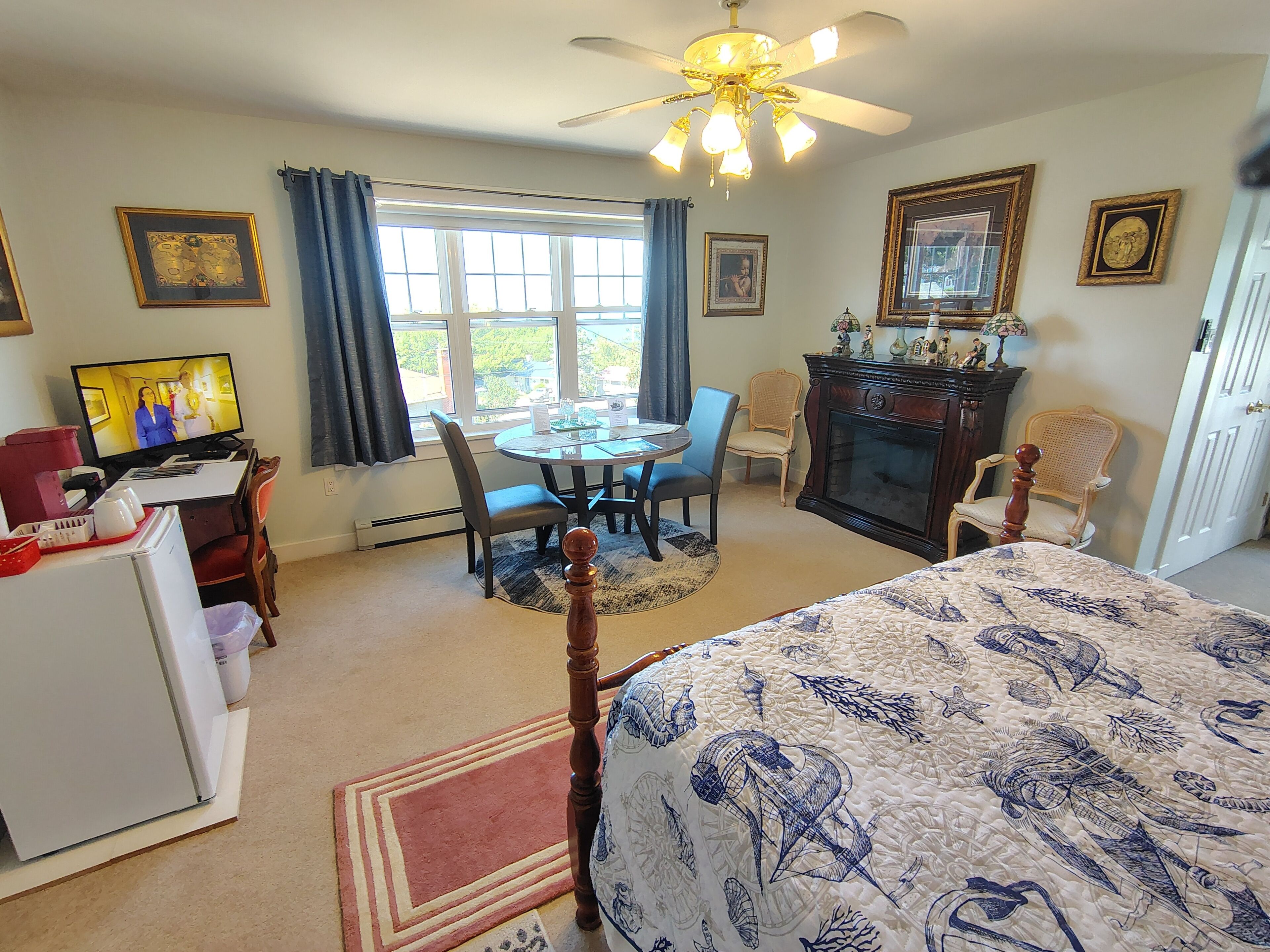 A room with a bed, a desk with a computer, a dining table with chairs, a fireplace, and a ceiling fan.