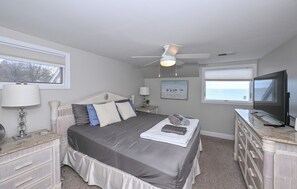 Un dormitorio con cama, ventilador de techo, televisor y ventana con vista al océano.