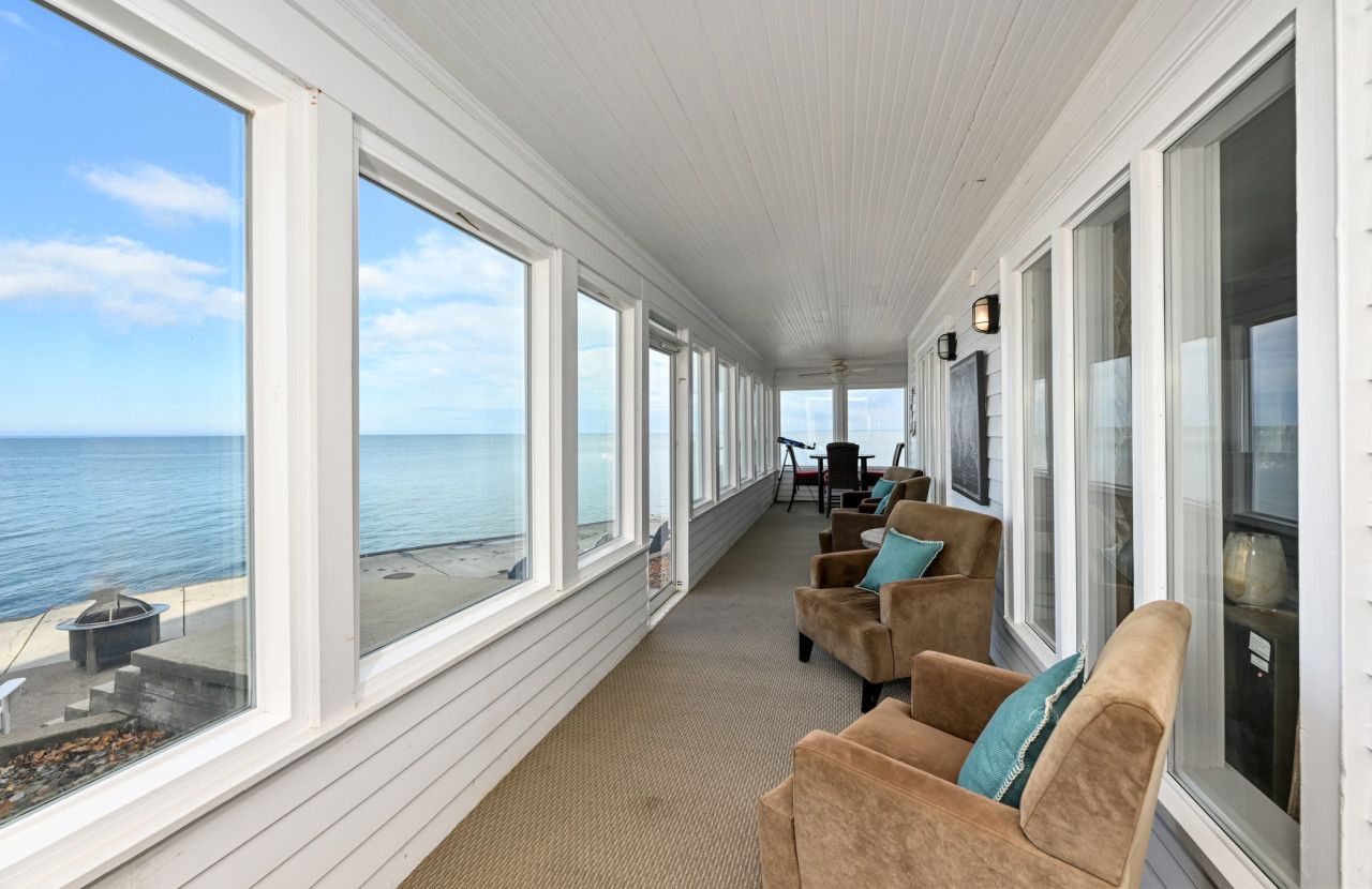 En overdækket veranda med store vinduer med udsigt over havet, møbleret med stole og et bord.