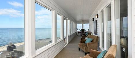 En overdækket veranda med store vinduer med udsigt over havet, møbleret med stole og et bord.