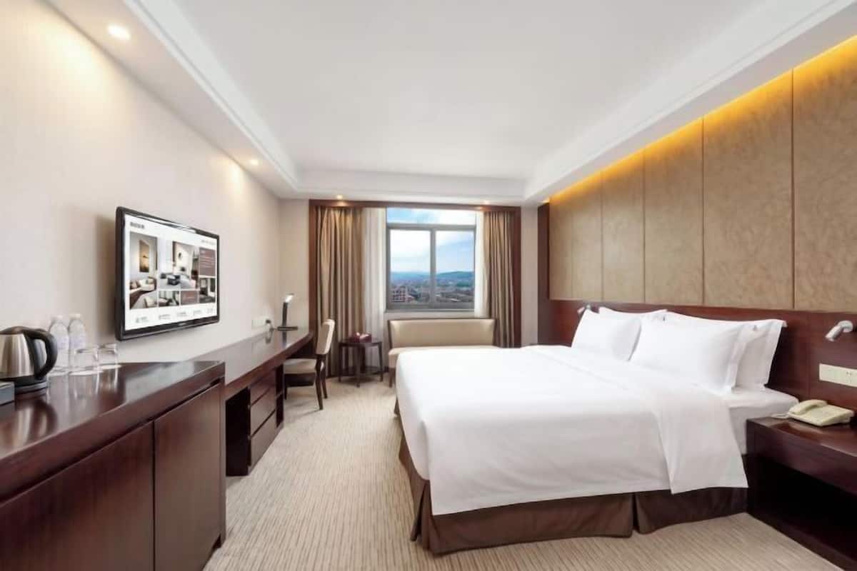 A hotel room with a bed, bedside table, desk, chair, and a large window with a view.