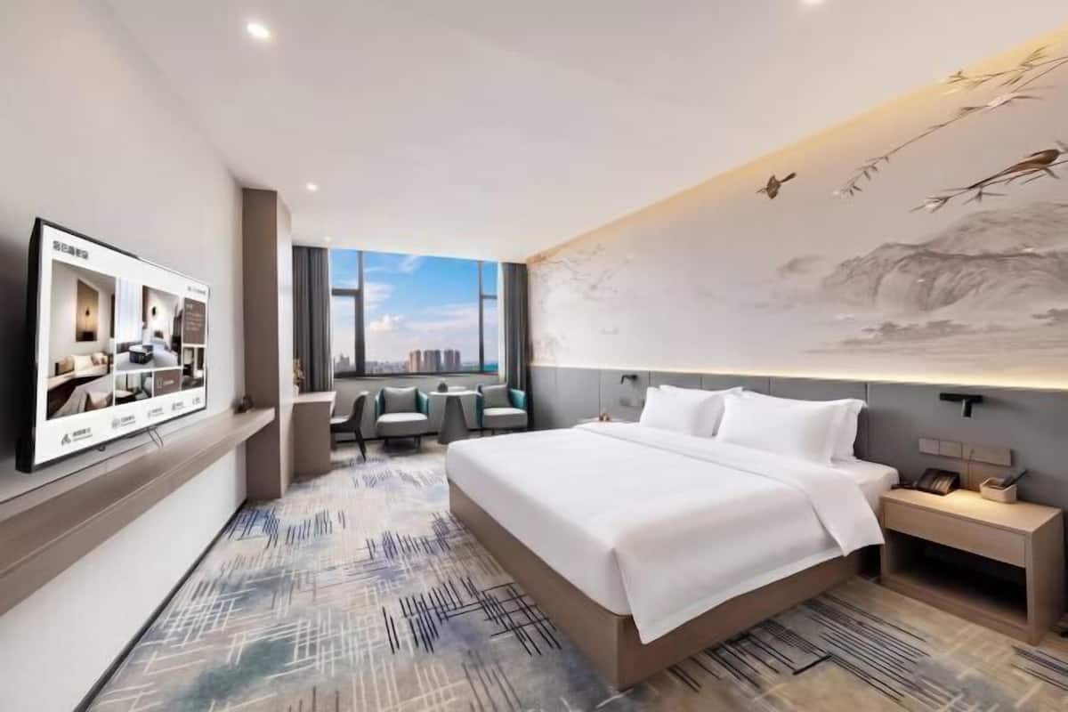 A modern hotel room with a large bed, a TV, a desk, and a view of the city.