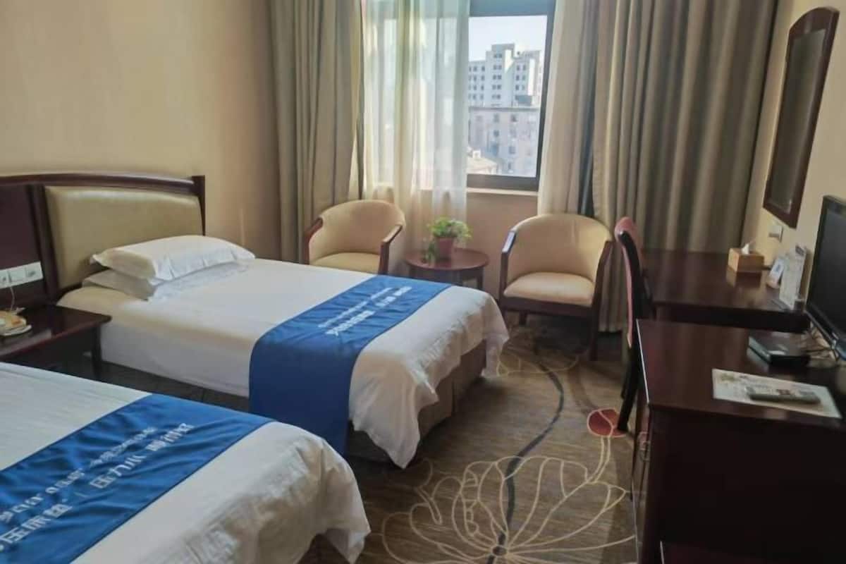 A hotel room with two beds, a desk, a chair, a small table with a plant, and a view of buildings through the window.