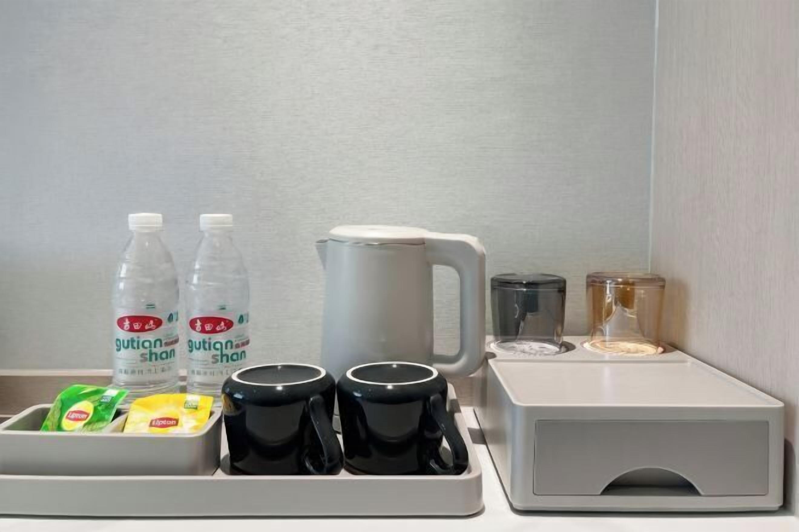 A hotel room's breakfast setup with a coffee maker, two bottles of water, a mug, a glass, and a tray with food items.