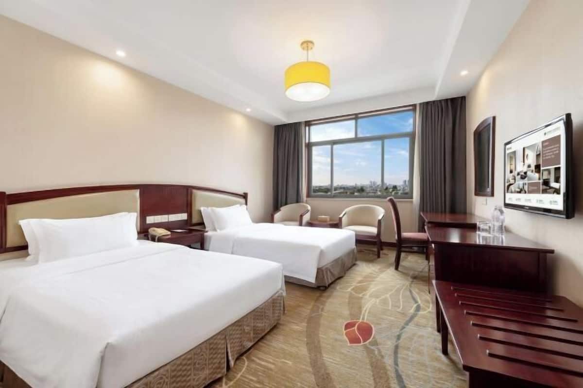 A hotel room with two beds, a desk, a chair, a large window with a city view, and a framed picture on the wall.