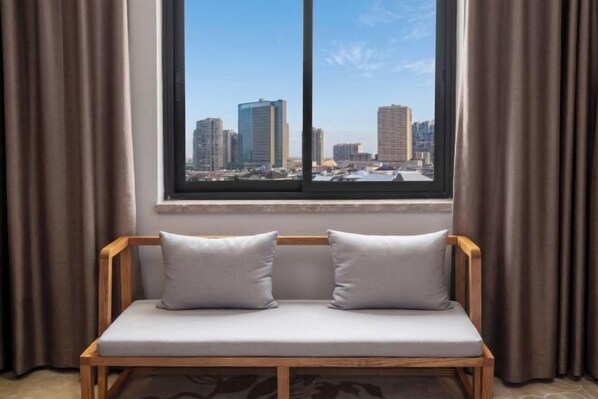 A wooden bench with two pillows, a window with a cityscape view, and brown curtains. - Lanjiang Hotel (Lanxi)