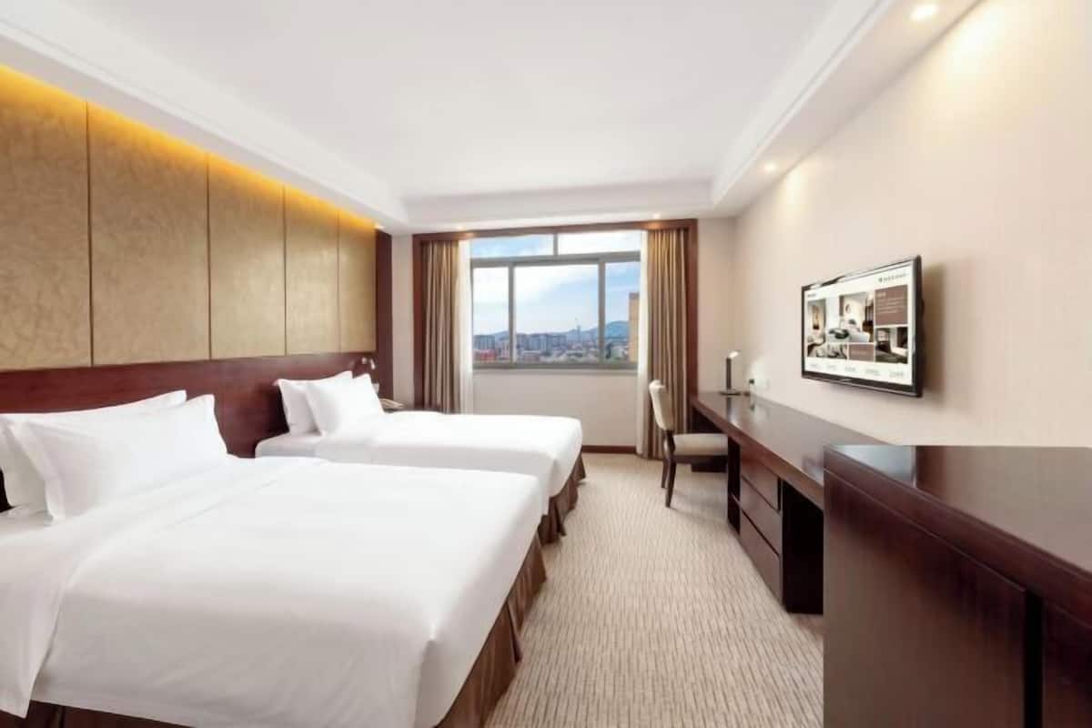 A hotel room with two beds, a desk, a chair, a TV, and a large window with a city view.