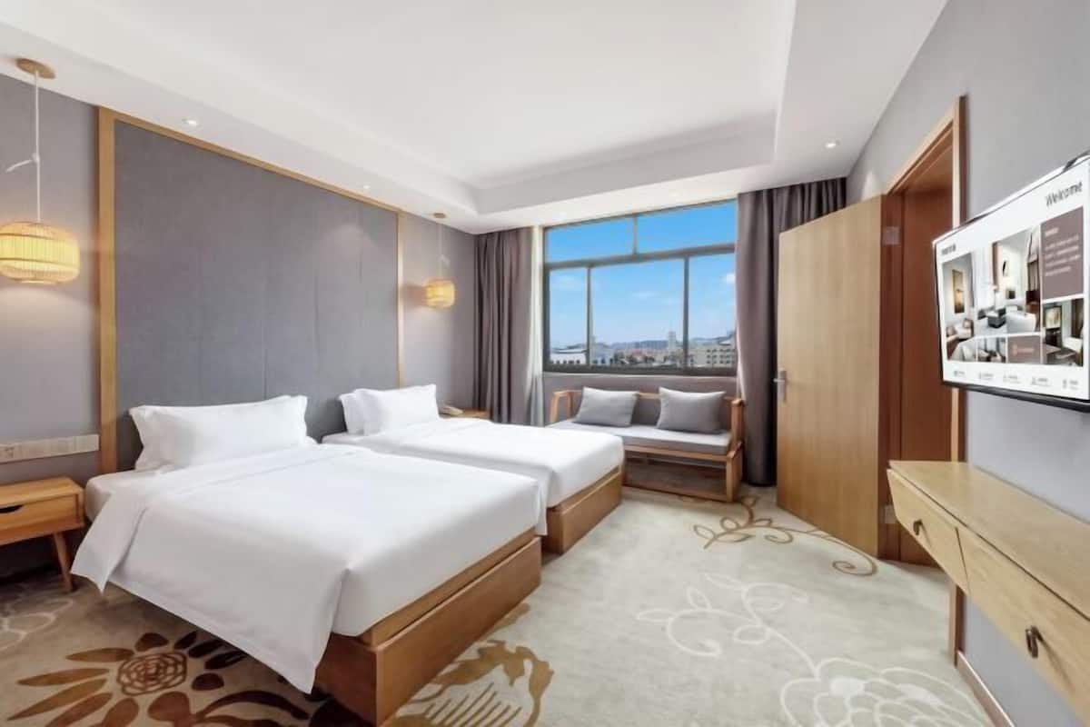 A hotel room with two beds, a desk, a TV, and a window with a city view.