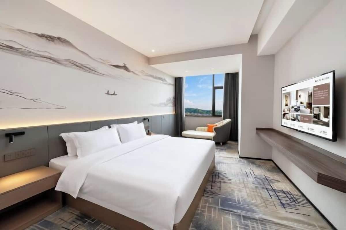 A modern hotel room with a large bed, a desk, a chair, a mounted TV, and a view of the outdoors.