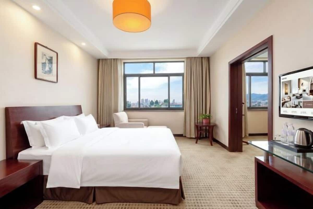 A hotel room with a bed, a chair, a desk, a window with a city view, and a door leading to another room.