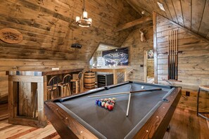 Un interior de madera con mesa de billar, sillas, zona de bar y un ciervo montado en la pared.