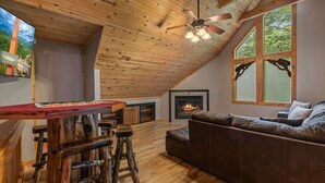 A loft-style room with a wooden ceiling, a fireplace, a sofa, and a dining area with stools. - Cabin for 12 | 10 mins to Pkwy w/ Hot Tub & Games! (Sevierville)