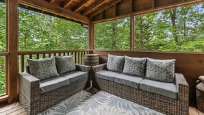 A covered outdoor seating area with wicker furniture and cushions, a wooden deck, and a view of trees. - Cabin for 12 | 10 mins to Pkwy w/ Hot Tub & Games! (Sevierville)