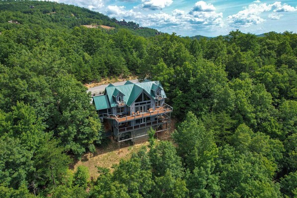 A large, multi-story house surrounded by dense forest, with a prominent green roof and multiple balconies. - 5BR Cabin w/ Mountain Views, Hot Tub & Game Room! (Sevierville)