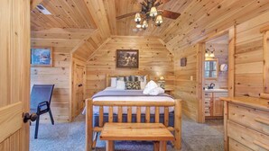 Camera da letto in una baita di legno, con un letto, una panca, una ventola a soffitto e un bagno piccolo.