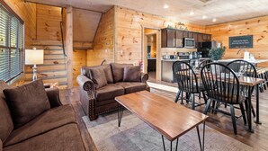 A cozy wooden interior with a sofa, dining table, and chairs, a kitchen area, and a staircase. - Modern Cabin w/ Pool, Hot Tub, Games & More! (Sevierville)