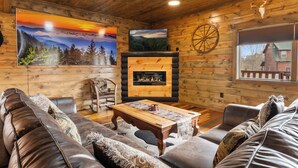 L’intérieur chaleureux d’un chalet avec une cheminée, des murs en bois, un grand tableau représentant un coucher de soleil et une vieille roue.