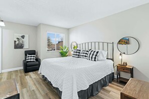 Quarto com cama, cadeira, mesa de cabeceira, luminária, janela e planta.