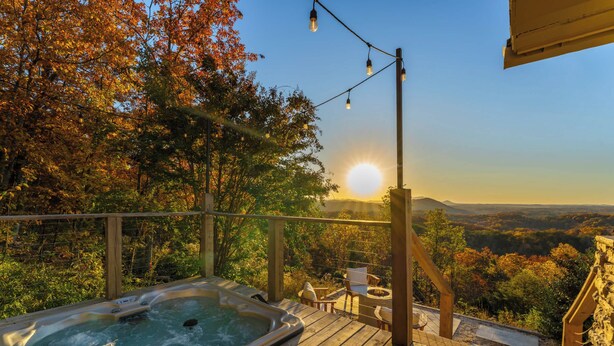 En træterrasse med jacuzzi, udendørs møbler og en smuk udsigt over skov og bjerge i solnedgangen.
