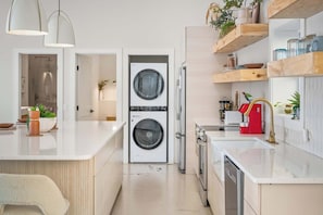 A compact kitchen with a built-in washer and dryer, a countertop with a sink, and wooden shelves above. - Modern Home w/ Hot Tub & Backyard Oasis! (Sevierville)