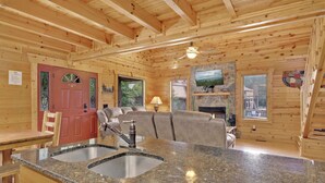 A wooden cabin interior with a kitchen area featuring granite countertops, a fireplace, and a television mounted above it.