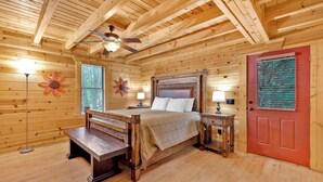 A wooden cabin bedroom with a bed, bedside tables, a bench, a ceiling fan, and a red door.