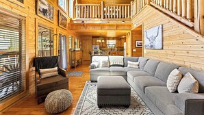 A spacious log cabin interior with a large gray sectional sofa, a wooden staircase, and a dining area. - Woodland Wonder w/ Hot Tub & Pool Table! Sleeps 8! (Sevierville)