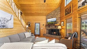 A cozy log cabin interior with a fireplace, a sofa, a chair, and a large window overlooking a scenic view. - Woodland Wonder w/ Hot Tub & Pool Table! Sleeps 8! (Sevierville)