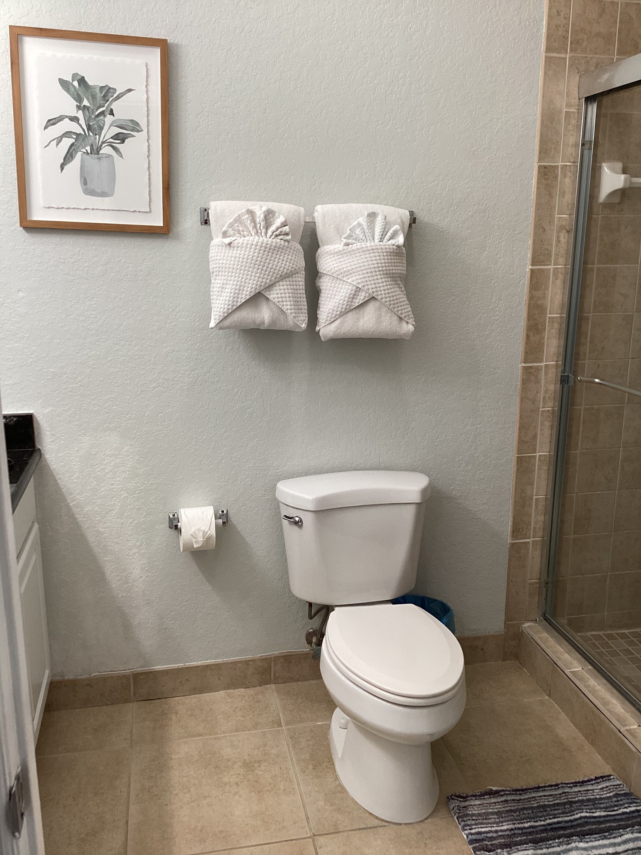 Een badkamer met toilet, twee handdoeken aan de muur, een lijst met afbeelding en een douchehoek.