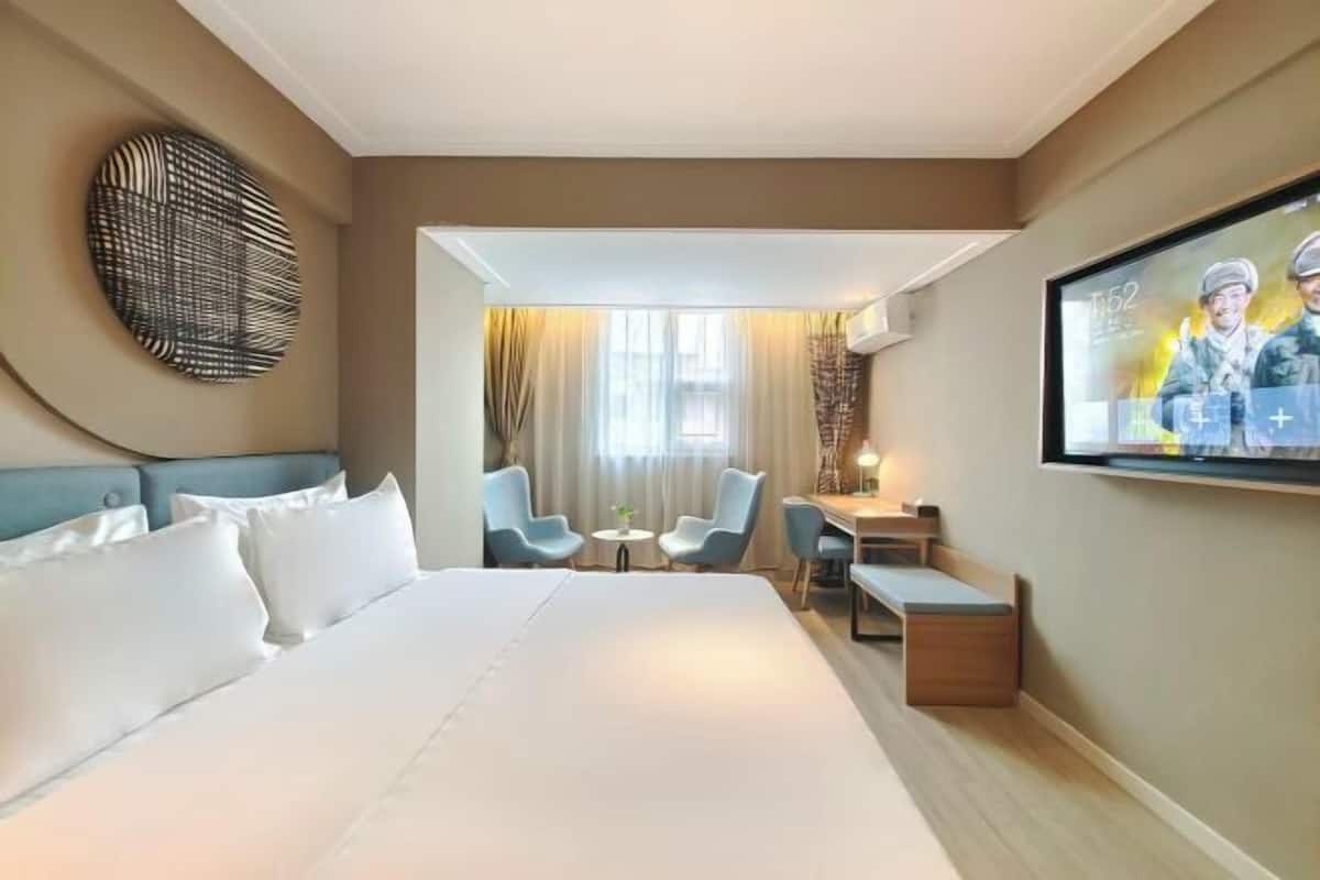 A hotel room with a large bed, a desk, two chairs, a TV, and a wall art piece.
