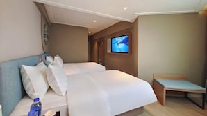 Et hotelværelse med en stor seng, et fjernsyn ophængt på væggen, en lille træstol og et natbord med en vandflaske og en kop.
