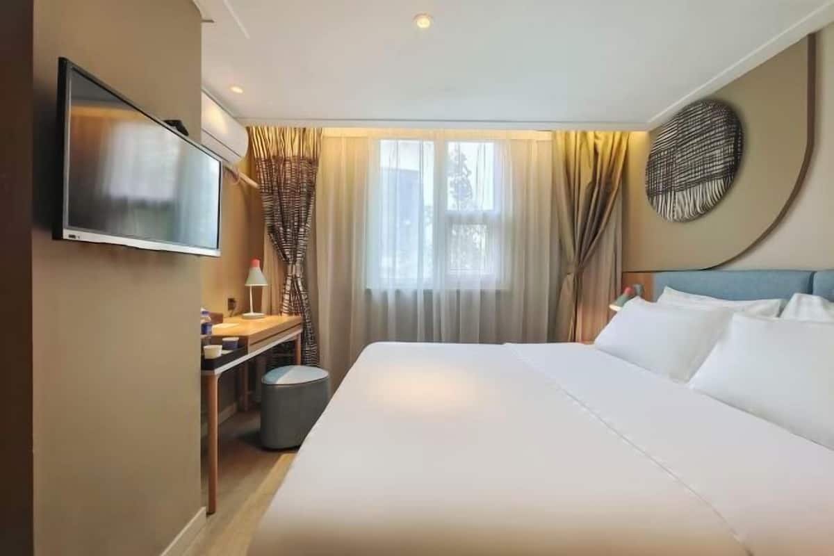 A hotel room with a bed, a TV mounted on the wall, a desk with a lamp, and a window with curtains.