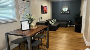 Ein modernes Wohnzimmer mit einem Holzesstisch, zwei Barhocker, einem dunklen Sofa und einem kleinen Couchtisch. Eine Karte ist an der Wand angebracht, und über dem Sofa hängt ein Spiegel.
