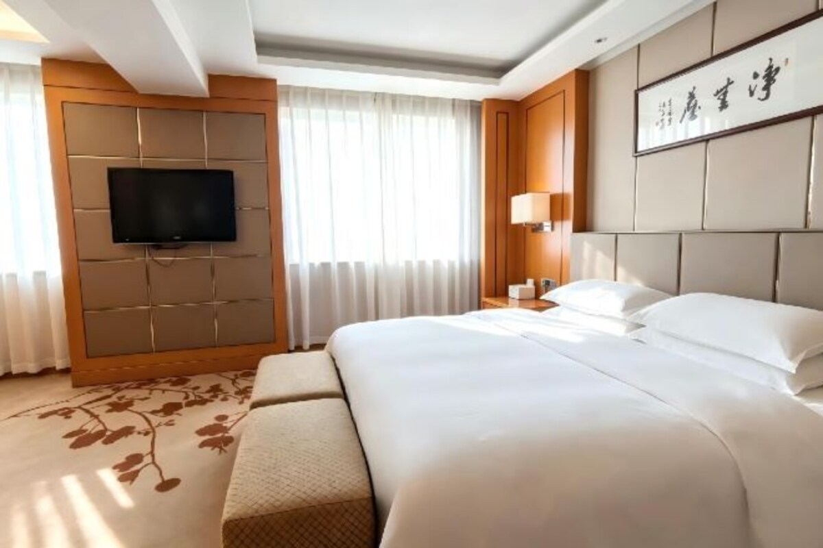 A hotel room with a large bed, a flat-screen TV mounted on the wall, a bedside table with a lamp, and a large window with curtains.