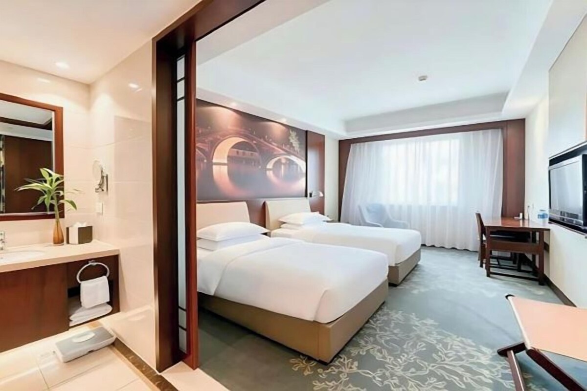 Habitación de hotel con cama, escritorio, silla, televisión y baño con espejo y lavamanos.