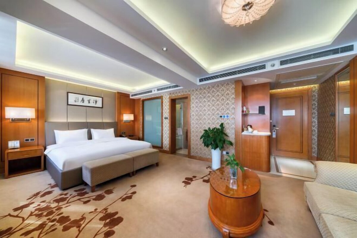 A modern hotel room with a large bed, a sofa, a round wooden coffee table, and a small desk.