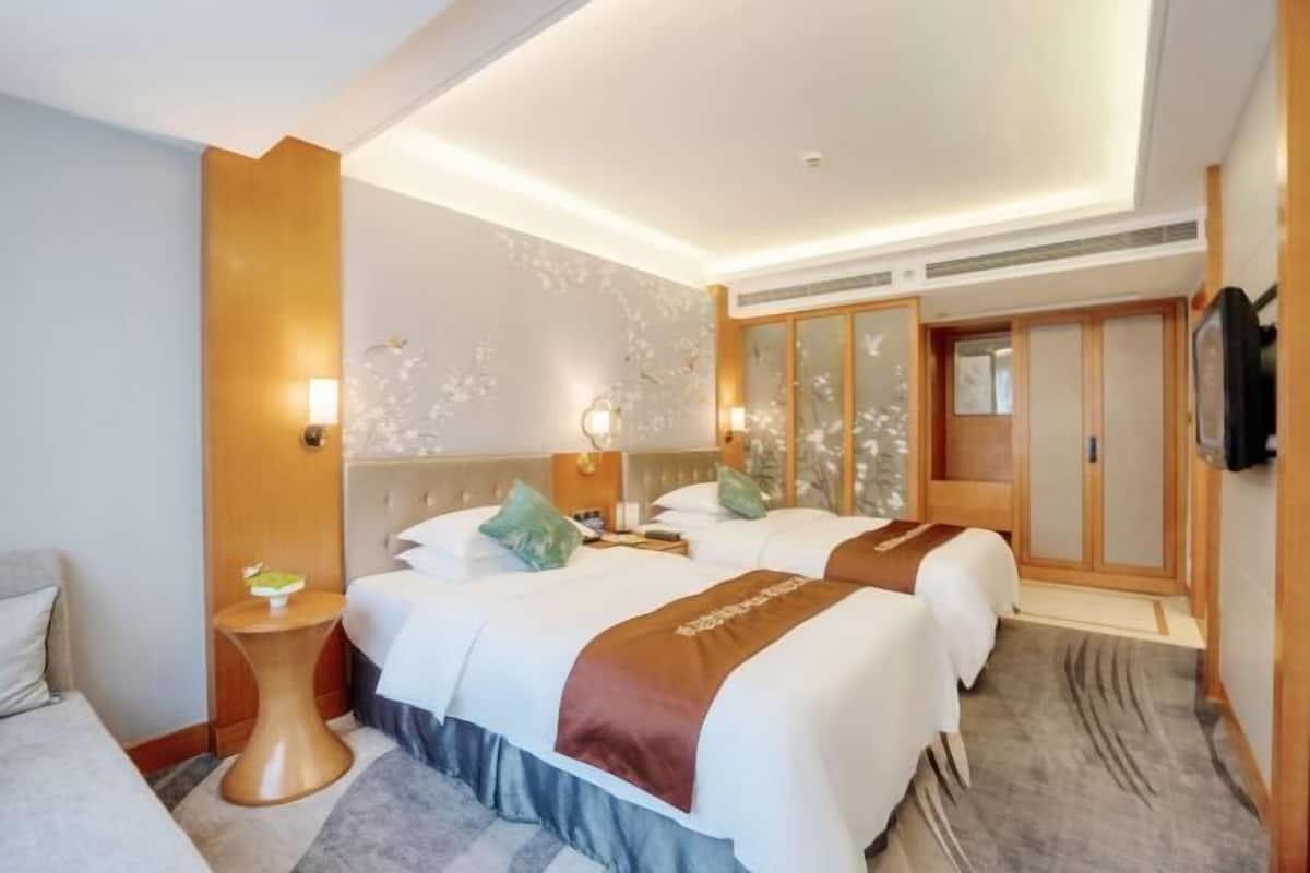 Una habitación de hotel con dos camas, un armario de madera, una mesita con una lámpara y un televisor en la pared.