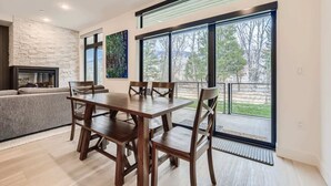 A modern living room with a dining table, chairs, a fireplace, and large windows offering a view of trees.
