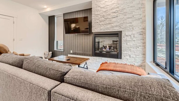 A modern living room with a sectional sofa, a flat-screen TV, a fireplace, and a large window.