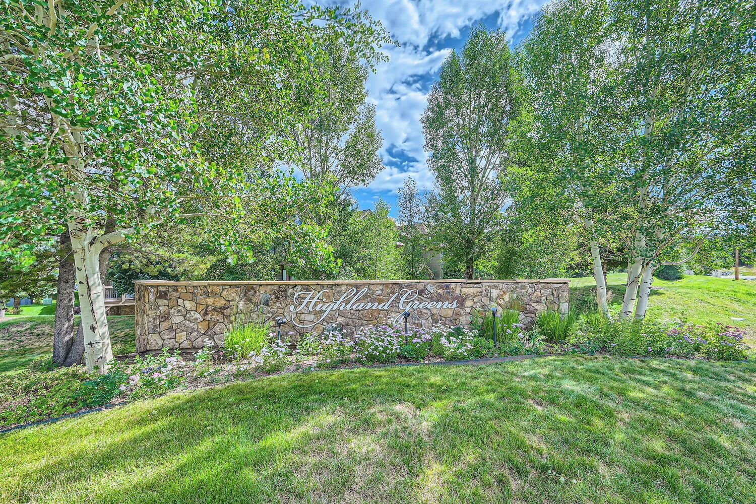 A stone wall with 'HIGHLAND GREENS' written on it, surrounded by lush greenery and trees.