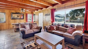 L’intérieur chaleureux d’un chalet avec des murs et un plafond en bois, un sol carrelé en pierre, un ensemble de canapés, une table à manger et une grande fenêtre donnant sur l’extérieur.