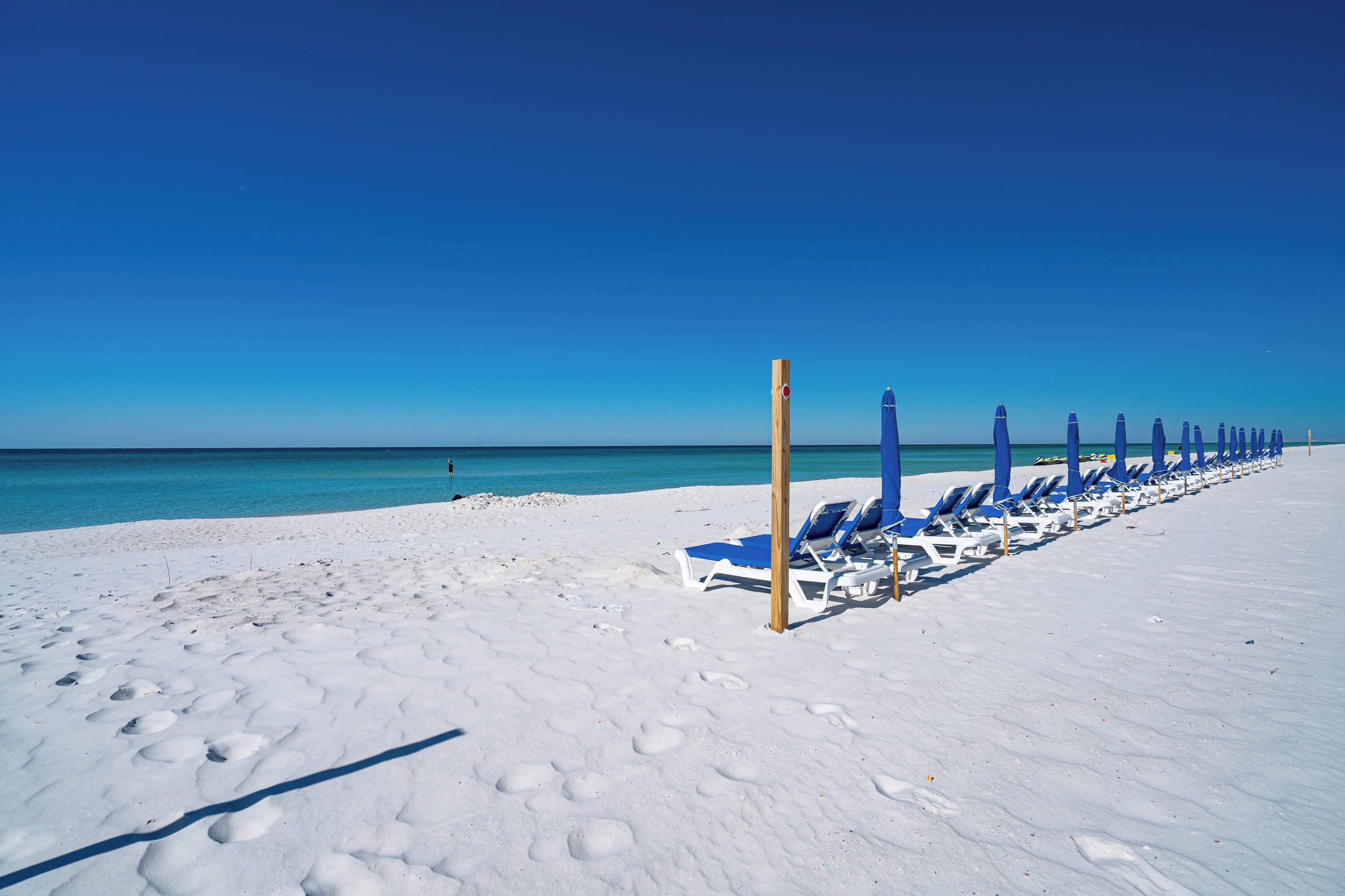 Una playa de arena blanca, una hilera de sillas azules para tomar el sol y un poste de madera.