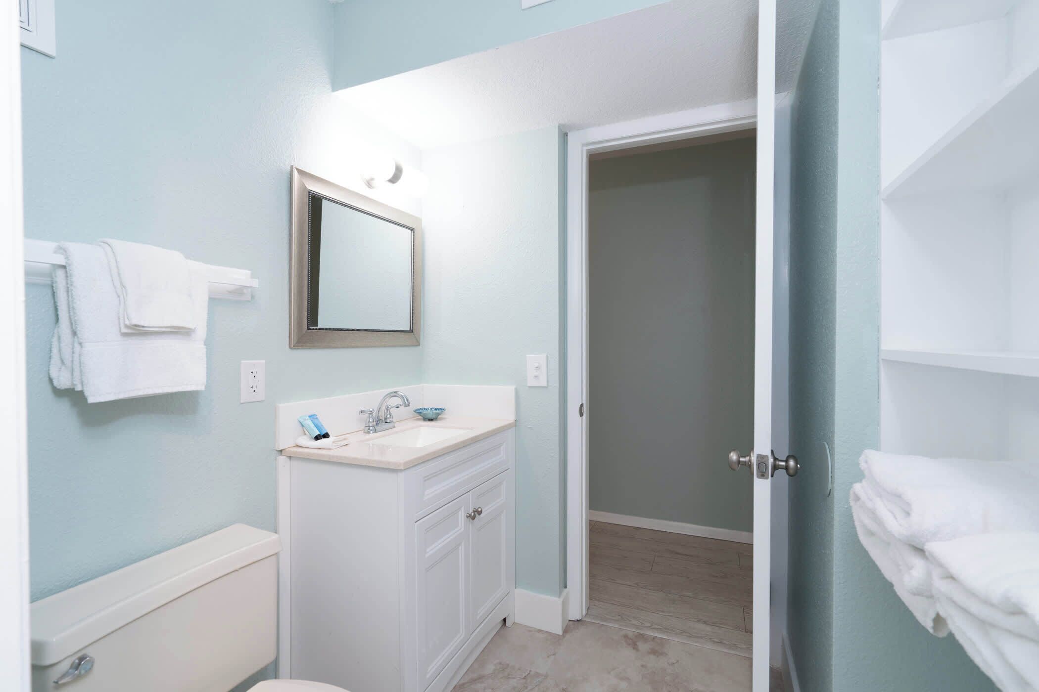 A bathroom with a white sink, mirror, and toilet. There are towels on a rack and a door leading to another room.